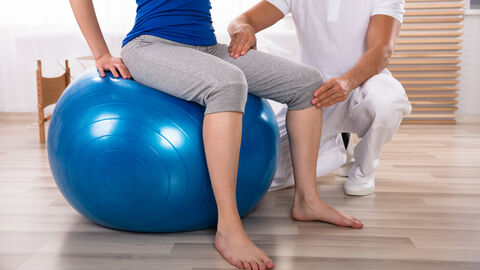Physiotherapeut trainiert mit einer Frau, während sie auf dem Fitnessball sitzt Physiotherapeut trainiert mit einer Frau, während sie auf dem Fitnessball sitzt